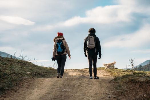 Young couple with backpacks and a dog walking in the mountains. Man and woman warmly dressed on hiking path. View from the back.