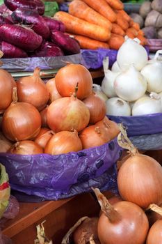 Various varieties of onions, yellow, white, red against the background of other vegetables, carrots, beets in blur are sold in trays on the market. Vertical image, close-up.