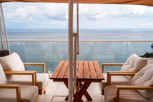 Restaurant with sea view. You can see rocks in the sea. Wooden tables and chairs with white cushions. Overcast, cloudy weather