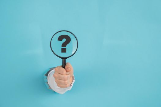 A woman's hand sticking out of a hole on a blue paper background holds a magnifying loupe with a question mark
