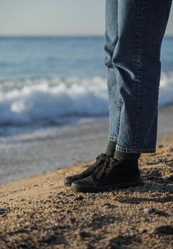 Legs in blue jeans and black shoes standing on the beach with sea waves at the background.