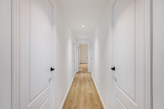 Interior of long narrow hallway with closed doors, wooden floor and white walls in apartment designed in minimal style.