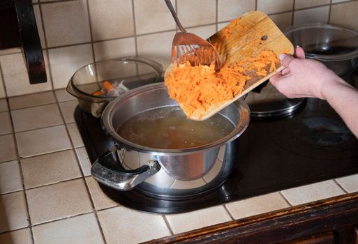 a woman puts grated carrots in a pan in the kitchen against the background of fresh vegetables, ingredients for step by step cooking soup. High quality photo