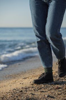 A woman in blue jeans and black shoes walking on the beach with sea waves at the background.