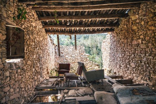 Abandoned stone house with wooden roof and broken wall with outside view. Ruin of ancient deserted building with old furniture. View from inside.