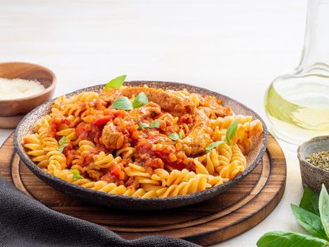 fusilli pasta with tomato sauce, chicken fillet with basil leaves on light white wooden background, side view.