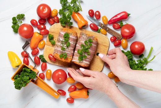 female hands decorate with parsley minced meat sausages in bacon with fresh vegetables, cherry tomatoes, sweet peppers, colorful food flat lay. High quality photo