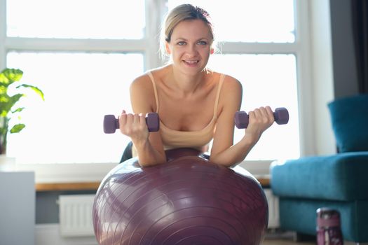 Portrait of smiling woman doing exercise with dumbbells on fitness ball. Fitness equipment for sport activity. Healthy lifestyle concept