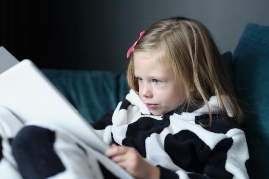 Portrait of little kid sitting on sofa and reading book. Distance education and home learning or leisure time at home