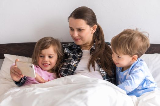 Happy Young Woman Mothefamilyr With 3-4 Aged Children Siblings Baby Boy And Girl Watching Smartphone Together Digital Tablet On White Bed. Mom With Daughter And Son Family Making Selfie In Pajamas