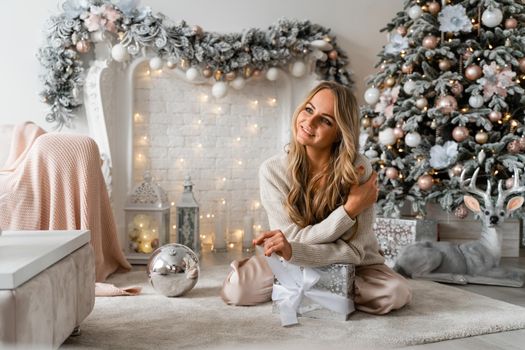 Happy blonde beauty enjoys Christmas Under the Christmas tree in a cozy home, holding a box with a gift