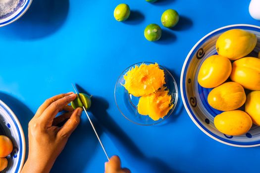 Top view of a latin woman's hand cutting a lemon on a blue surface with jocote mangoes and traditional summer fruits in Nicaragua, Central America and Latin America.