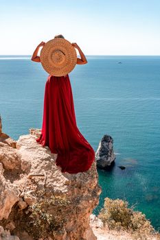 A girl with loose hair in a long red dress descends the stairs between the yellow rocks overlooking the sea. A rock can be seen in the sea. Sunny path on the sea from the rising sun.