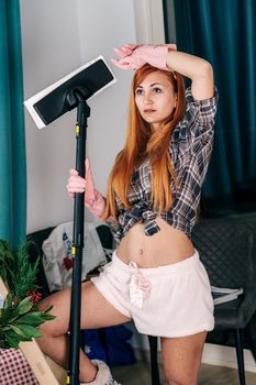 A young housewife in rubber gloves and a shirt holds a vacuum cleaner in her hands. Tired girl