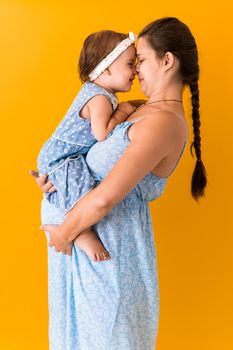 Motherhood, hot summer - portrait young pregnant happy smiling joy mother woman in blue dress holding little baby daughter toddler sibling bare feet in arms looking into eyes kiss on yellow background.