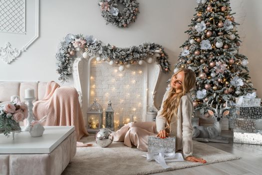 Happy blonde beauty enjoys Christmas Under the Christmas tree in a cozy home, holding a box with a gift