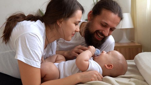 Woman And Man Holding Newborn. Mom, Dad And Baby On Bed. Close-up. Portrait of Young Smiling Family With Newborn On Hands. Happy Marriage Couple On Background. Childhood, Parenthood, Infants Concept.