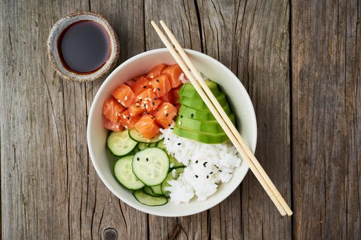 Salmon poke bowl with fresh fish, rice, cucumber, avocado with black and white sesame. Old wooden table. Food concept.