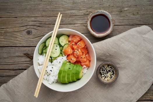 Salmon poke bowl with fresh fish, rice, cucumber, avocado with black and white sesame. Old wooden table. Food concept.
