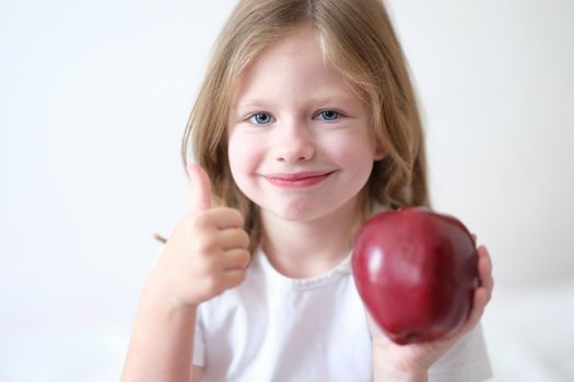 Portrait of little girl holding red apple in hands and showing thumbs up gesture. Healthy food and vitamins