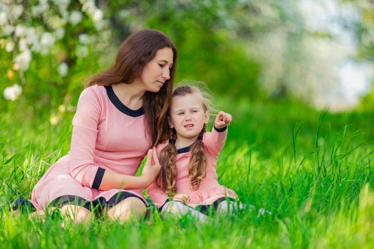 mother and daughter sit in the green grass against the backdrop of blooming apple trees
