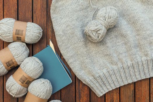 Top view of grey knitting yarns, notebook, pencil and a part of knitted sweater on brown wooden background. Hobby and needlecraft at home