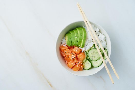 Salmon poke bowl with fresh fish, rice, cucumber, avocado with black and white sesame. White marble table. Food concept.