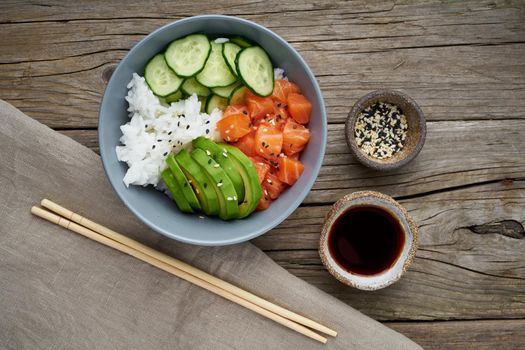 Salmon poke bowl with fresh fish, rice, cucumber, avocado with black and white sesame. Old wooden table. Food concept.