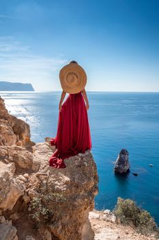 A girl with loose hair in a long red dress descends the stairs between the yellow rocks overlooking the sea. A rock can be seen in the sea. Sunny path on the sea from the rising sun.