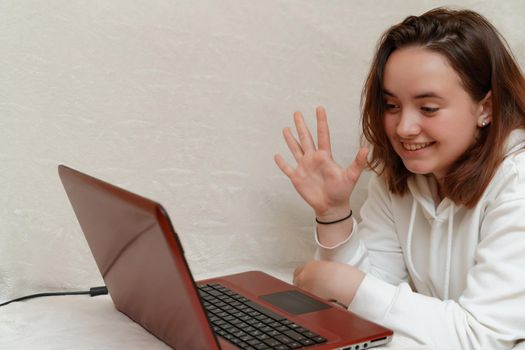 A young girl lies at home on the sofa with a computer and communicates via internet with friends, making hand gestures. covid-19 epidemic concept. Quarantine stay at home.