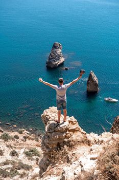 A male traveler in shorts and a T-shirt, facing the sea, raised his hands in the air. The concept of tourism and travel around the world