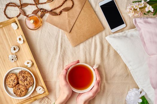 Woman holding mug of hot tea. Lazy cozy morning in bed. Flat lay woman accessories with letter, envelope, smartphone, perfume