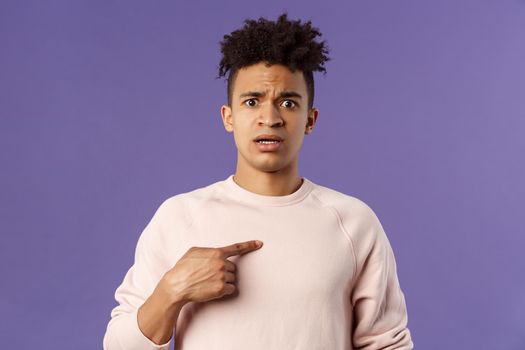 Close-up portrait of embarrassed and worried young man being named or called to principal office, pointing at himself and asking why with disbelief and anxiety, standing purple background.