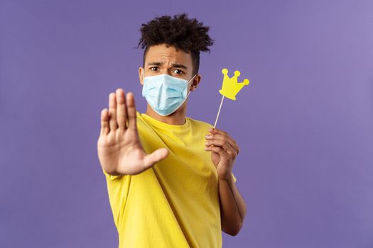 Close-up portrait of young hipster guy in medical mask stretch hand forward stop gesture, holding small paper crown, demand stay away, keep 1.5 meter distance, avoid social contact during quarantine.