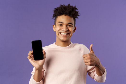 Close-up portrait of cheerful, satisfied young male customer recommend buying subscribtion to game or app, learning leanguage via application mobile phone, show thumb-up in approval.