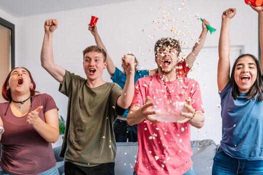 young friends excitedly throwing popcorn in the air after their team's victory, broadcast on television. group of young people watching football at home. leisure concept. happy and cheerful. natural light in the living room at home. trumpet