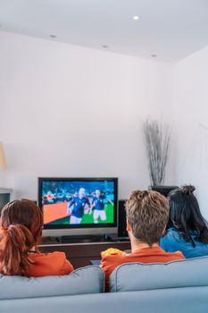 backs of friends cheering for their softball team while watching the game on TV on their living room couch. leisure concept, three young adults in blue t-shirts. happy and cheerful. natural light in the living room at home. trumpet
