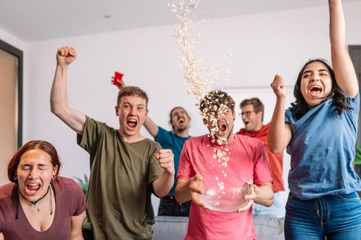 friends jumping for joy, throwing popcorn in the air after their team's victory. group of young people watching football at home. leisure concept. happy and cheerful. natural light in the living room at home. trumpet