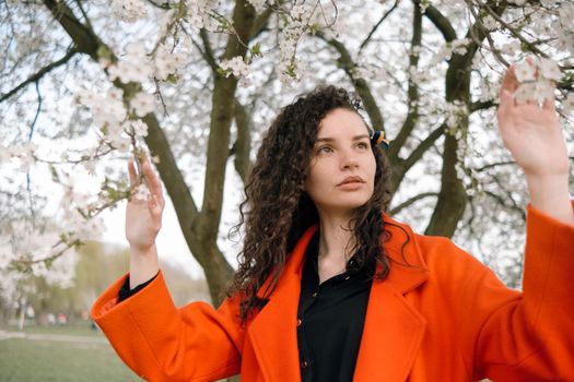 portrait of curly brunette woman in red coat in the park near flowering tree with ukrainian symbol in hair
