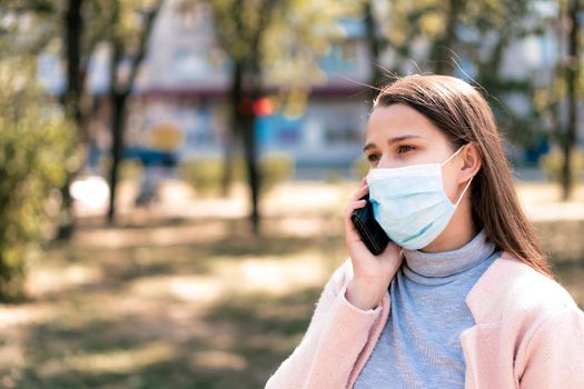 care, infants, spring, cvid-19, quarantine concept - Young cute long haired woman European Caucasian Slavic appearance in medical protective mask talking on phone in midday sunlight backlight in park.