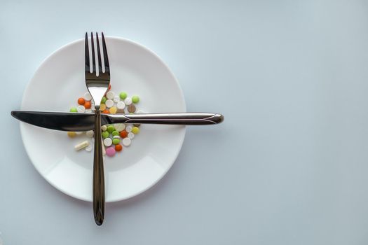 Pills on a white plate. Gestures with a knife and fork on a plate with pills. Medicines on a white background. A pile of multi-colored tablets disguised as food on a plate with cutlery..