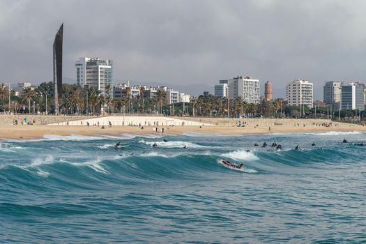 Many surfers at blue sea waves on beach with the background of Barcelona city on sunny day