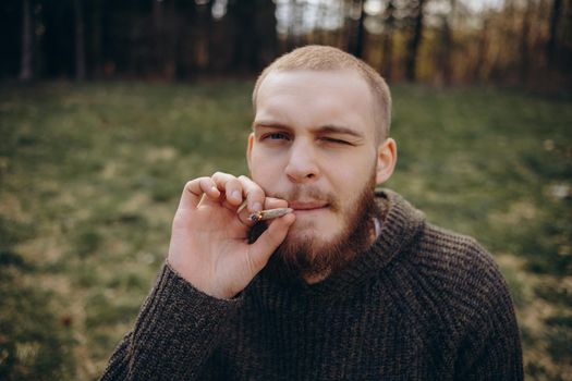 Young man beard smokes cigarette or tobacco. Guy is addicted to cigarettes and always smokes lot of tobacco every day. He smokes cigarette outdoors on background of lake in park. Cool guy blows smoke