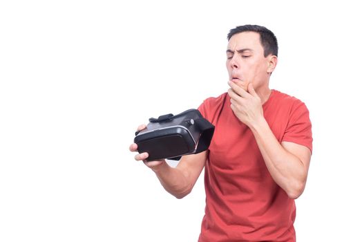Bewildered male gamer with hand at chin looking at modern VR headset with confuse while standing isolated on white background in light studio