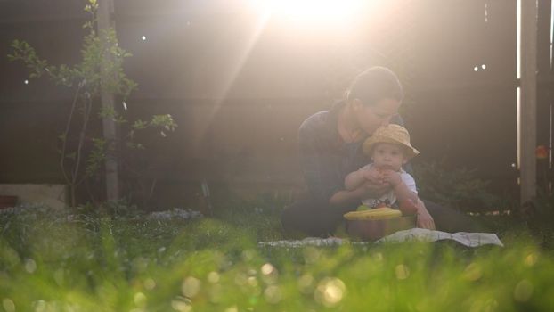 Happy Young Cheerful Mother Holding Baby Eating Fruits On Green Grass. Mom Adorable Infant Child Playing Outdoors With Love In Backyard Garden. Little Kid With Parents. Family, Nature, Ecology Concept.