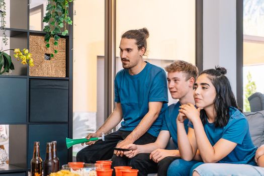 group of friends looking forward to the possible victory of their sports team, broadcast on television. leisure concept. three young adults in blue jerseys and red jerseys. happy and cheerful. natural light in living room at home.