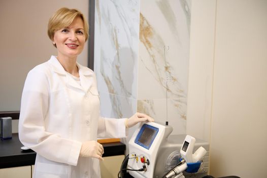 Experienced charming Caucasian mature female doctor in white medical gown and sterile surgical gloves , smiles at camera standing near ultrasound medical equipment in doctor's cabinet