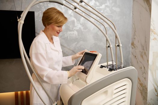 Confident doctor cosmetologist adjusting programs on the monitor of a modern ultrasound medical equipment before treating patient in a clinic specializing in hardware cosmetology