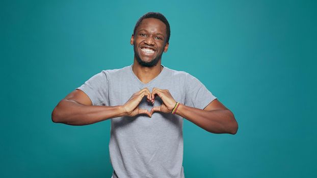 Smiling man making heart shape sign with hands in front of camera, showing romantic gesture of love and affection. Affectionate person doing romance and fondness symbol to express feelings.