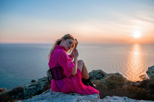 Side view a Young beautiful sensual woman in a red long dress posing on a volcanic rock high above the sea during sunset. Girl on the nature on overcast sky background. Fashion photo